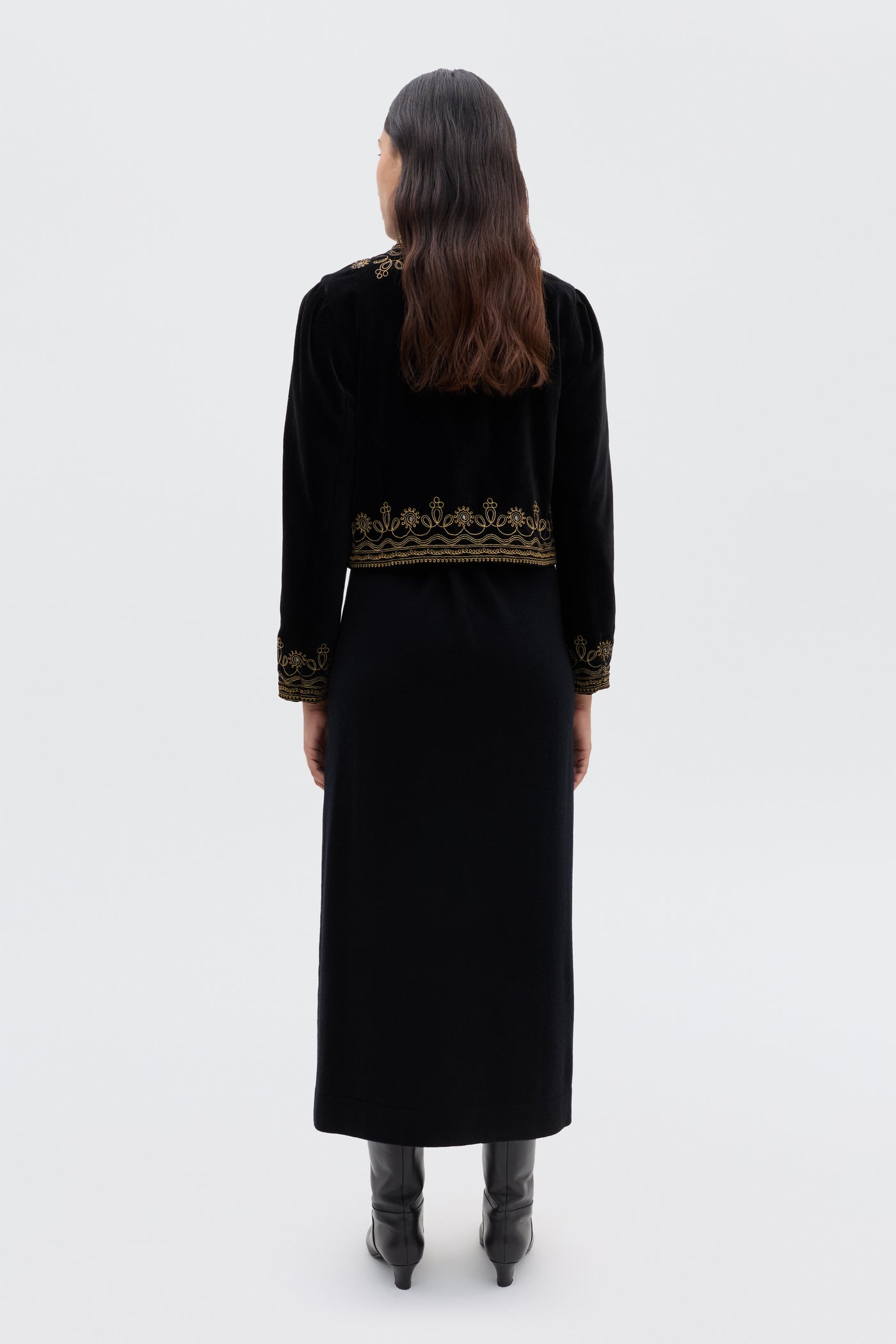 A woman with long brown hair, facing away, wears the Denver - Soutache Black: a black velvet jacket with gold embroidery on the hem and cuffs, over a long black dress and boots. The background is plain light gray.