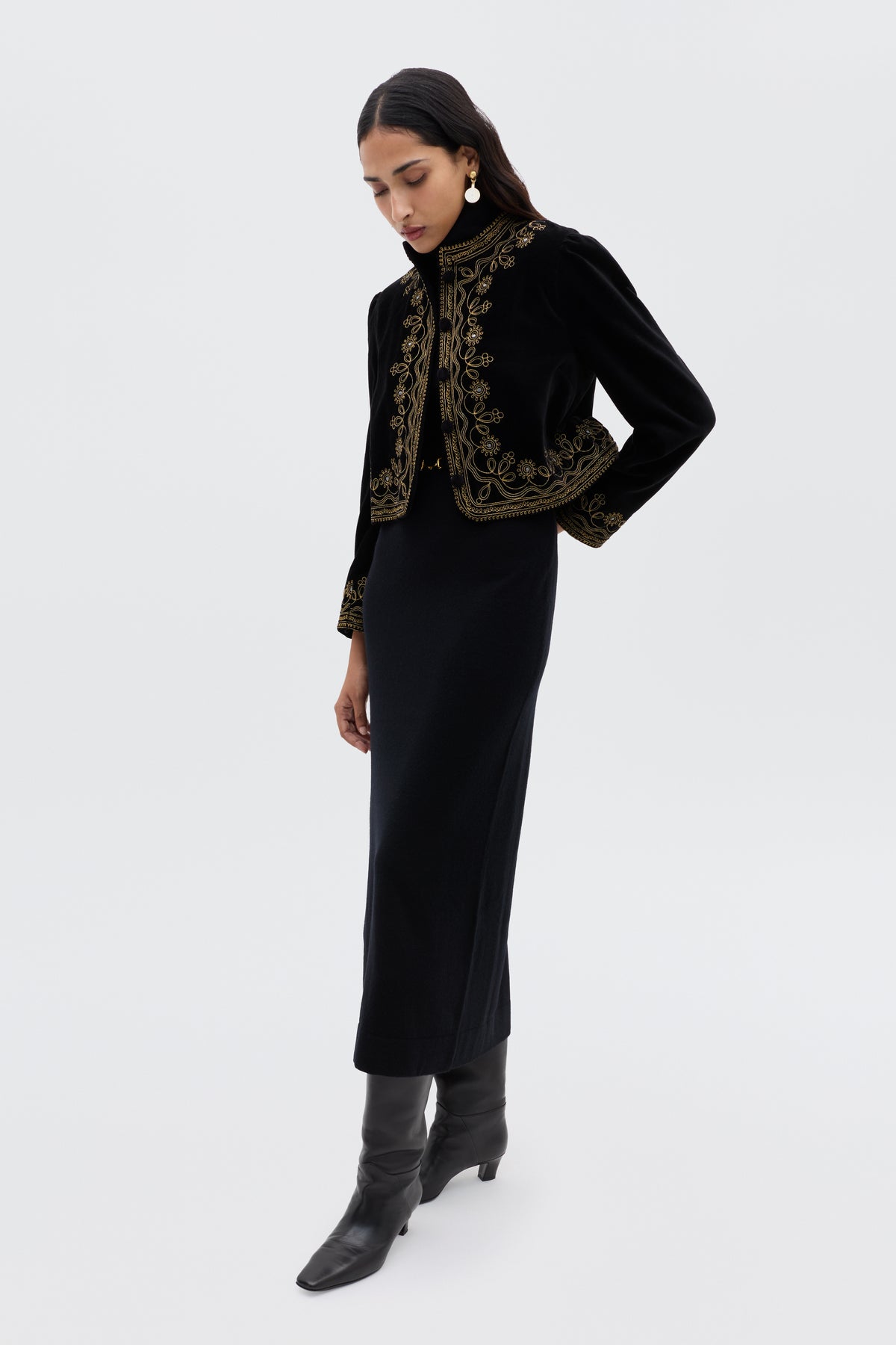 A woman stands against a plain white background wearing the Denver - Soutache Black, a long black dress paired with a black velvet jacket with gold embroidery, black knee-high boots, and gold earrings.