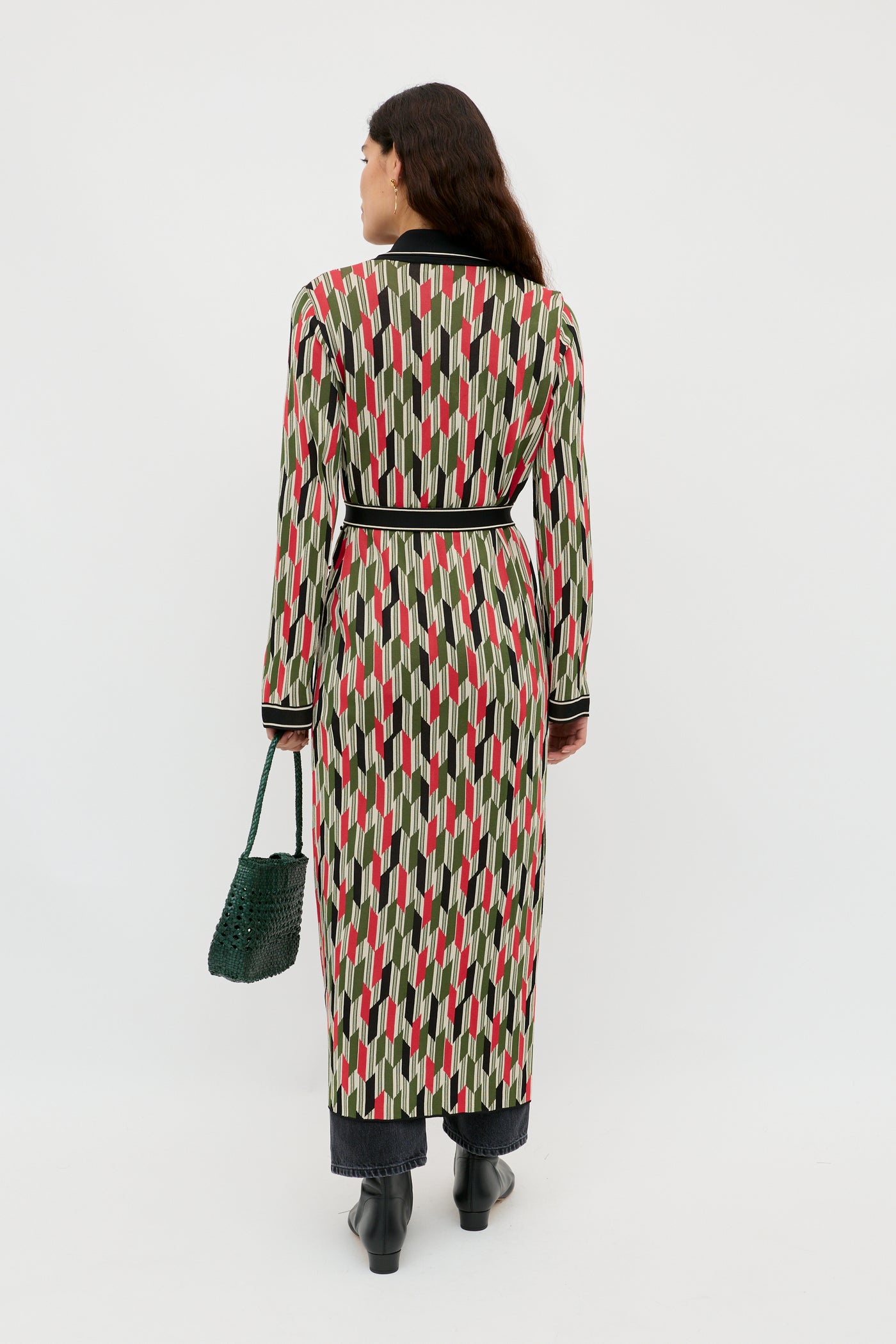 A woman faces away wearing the Apollo - Chevron Yarn Dyed Cream duster with red, black, and beige geometric shapes, paired with black pants and boots. She holds a green textured handbag against a plain white background.