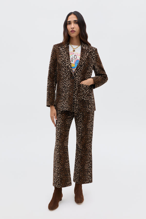 A woman with long, dark hair and a neutral expression wears the Xander - Lyra Leopard Black jacket, paired with flared pants, a graphic tee, brown ankle boots, and layered necklaces, standing against a plain light background.