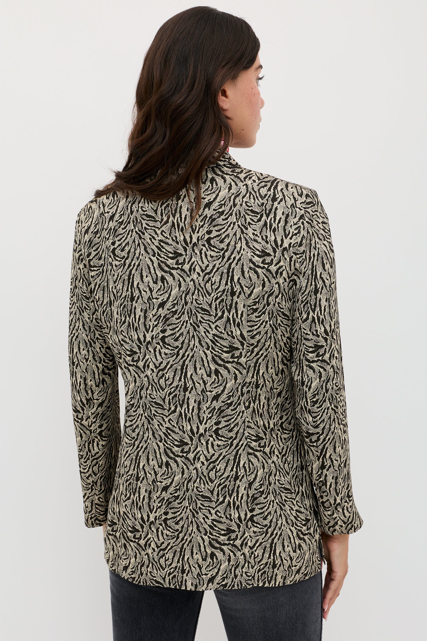 A person with wavy, dark brown hair stands facing away against a plain white background, wearing the Marselle - Sun Leo Tan long-sleeved black and beige animal print blazer.
