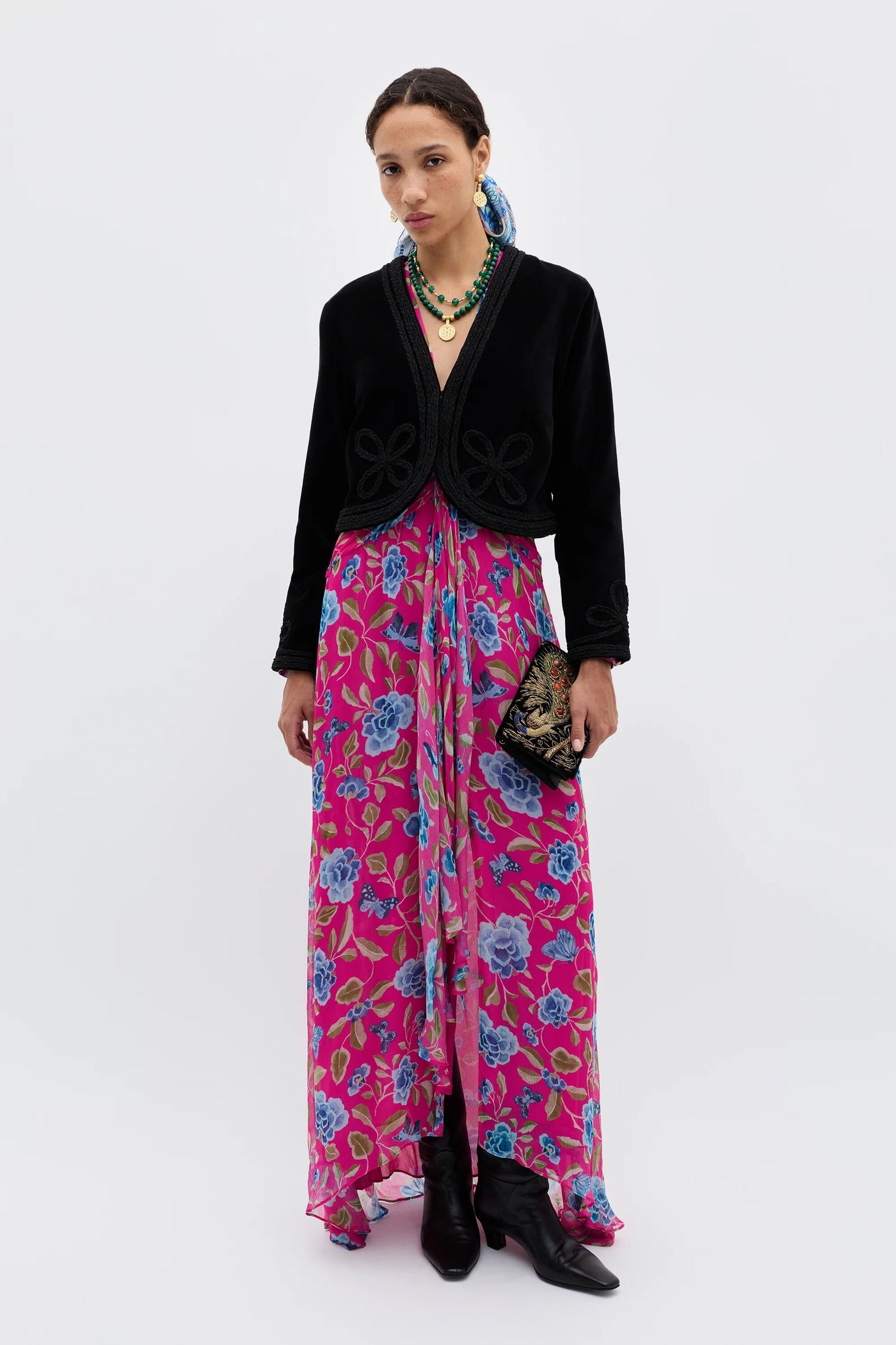 A woman stands against a plain background wearing the Kesia - Clover Appliqué Black jacket over a bright pink floral dress, paired with black boots, layered necklaces, and her hair styled back as she holds a decorative clutch.