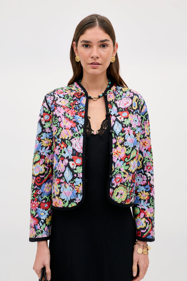 A woman wears the Lyanne - Ikat Bloom Charcoal jacket, featuring a colorful floral pattern over a black dress, accessorized with gold earrings, a necklace, and bracelets, standing against a plain light background.