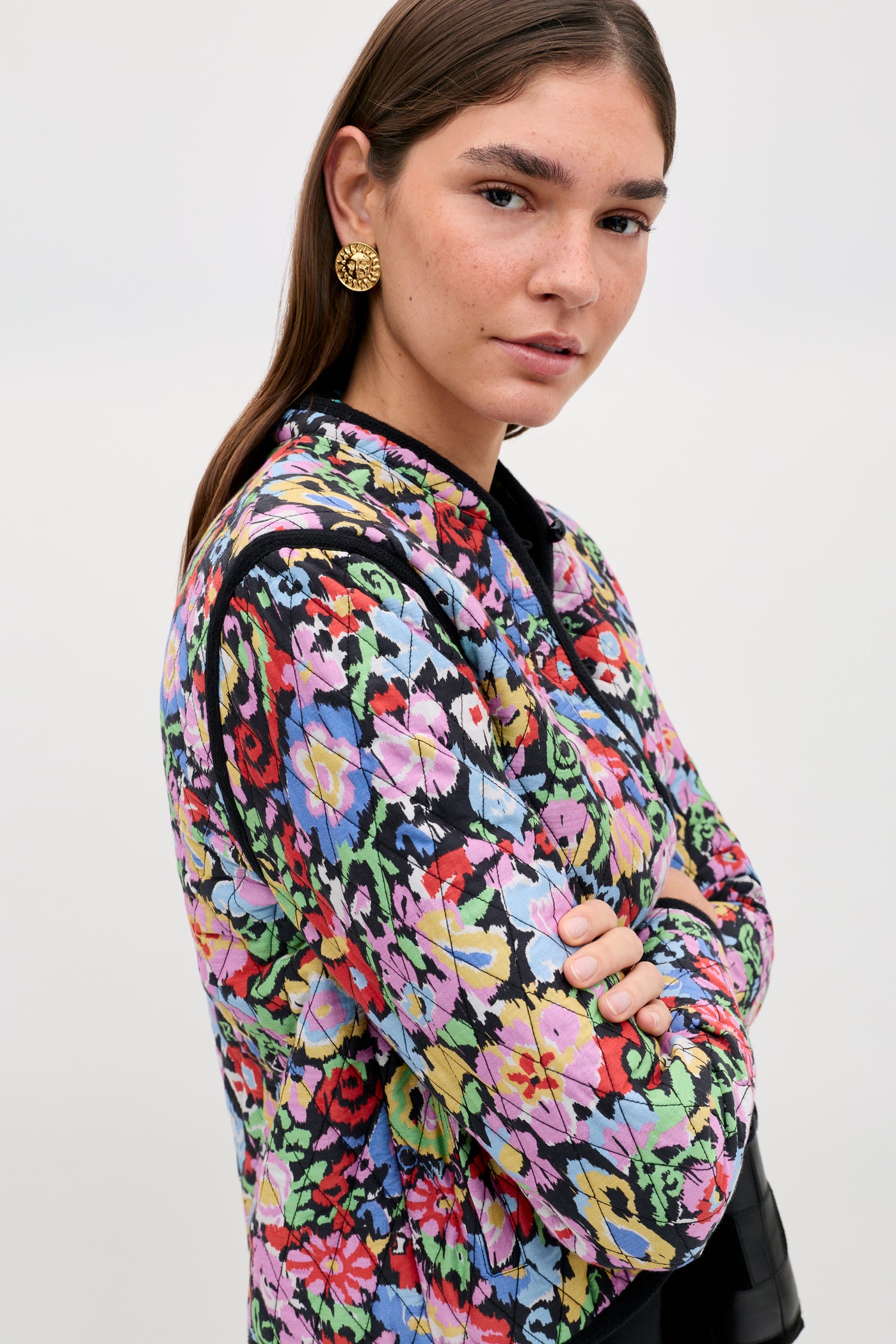A woman with straight brown hair wears gold earrings and the Lyanne - Ikat Bloom Charcoal jacket, featuring a colorful floral pattern. She stands with her arms crossed, glancing slightly over her shoulder against a plain light background.