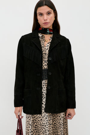 A woman stands against a plain background wearing the Teagan - Black suede jacket with fringe detailing over a leopard print dress, accessorized with a red floral scarf and holding a small textured bag.