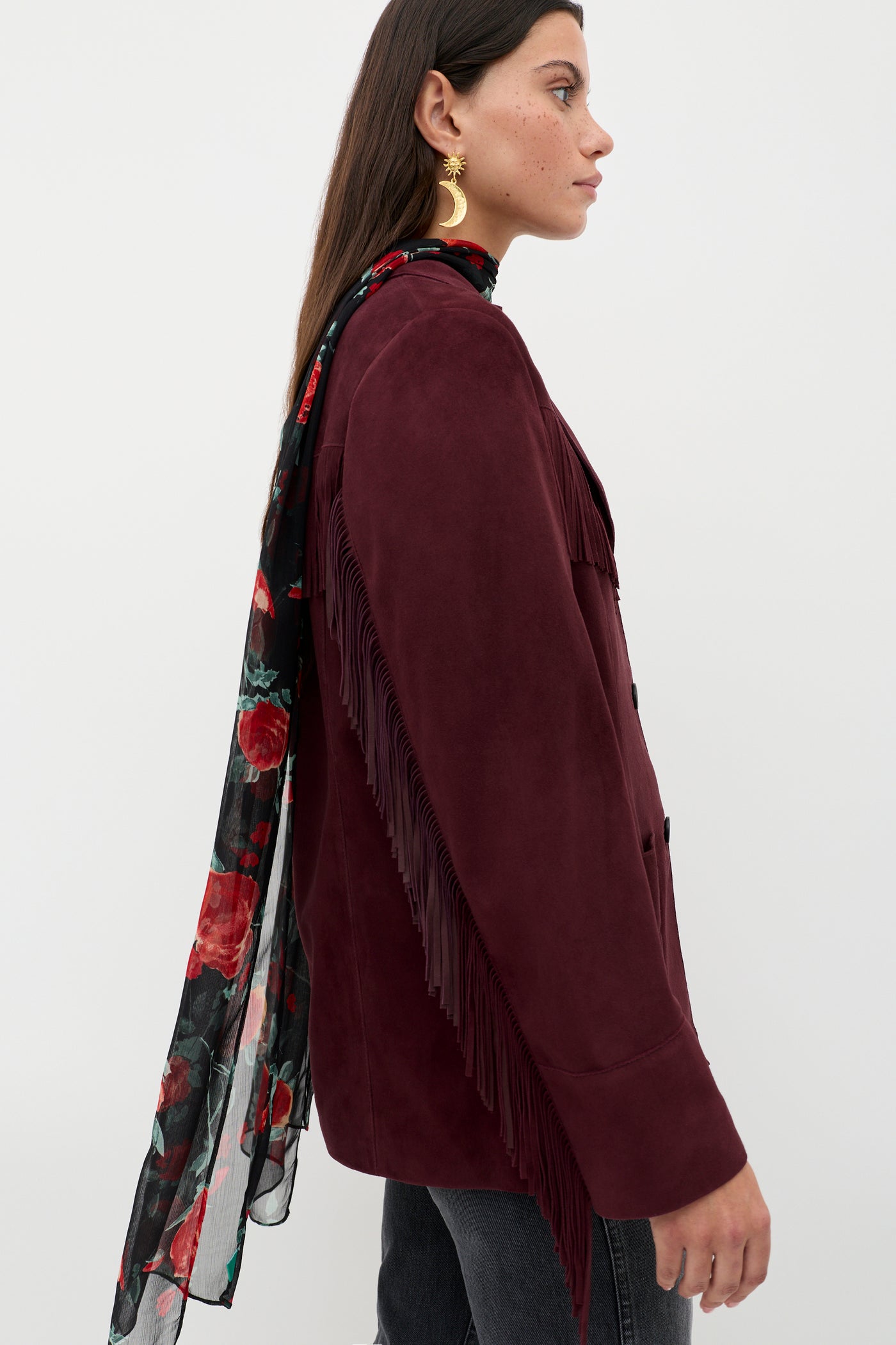 A woman in profile wears the Teagan – Maroon fringed jacket with dark jeans, gold earrings, and a long sheer black scarf with red floral designs. Her straight brown hair is worn down.