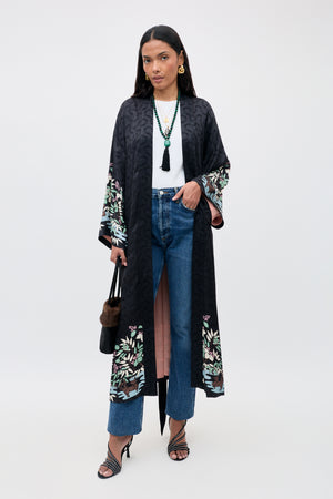 Wearing the Kaiden - Blossom Jungle Black kimono over a white top and blue jeans, she completes her look with strappy heels, a black handbag, layered necklaces, and straight dark hair.