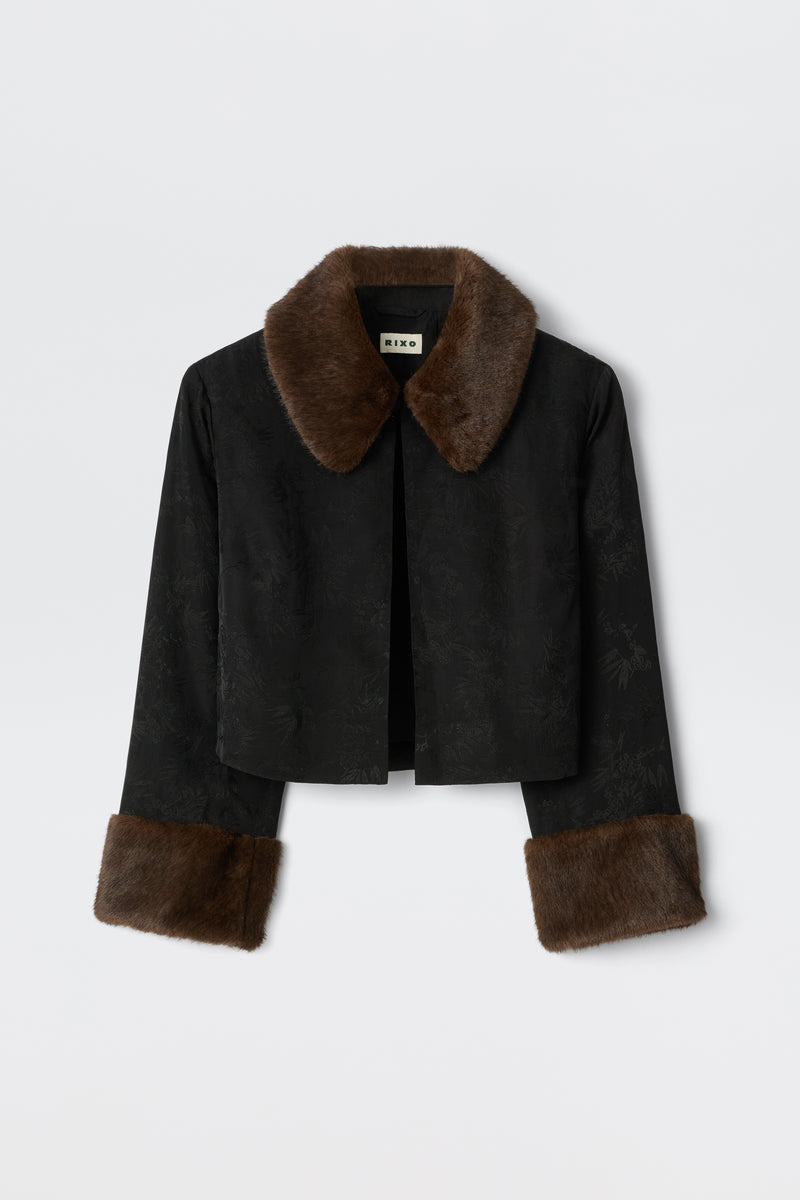 The Luanne - Black is a cropped jacket with a subtle floral pattern, wide brown faux fur collar, and matching cuffs, shown against a plain white background.