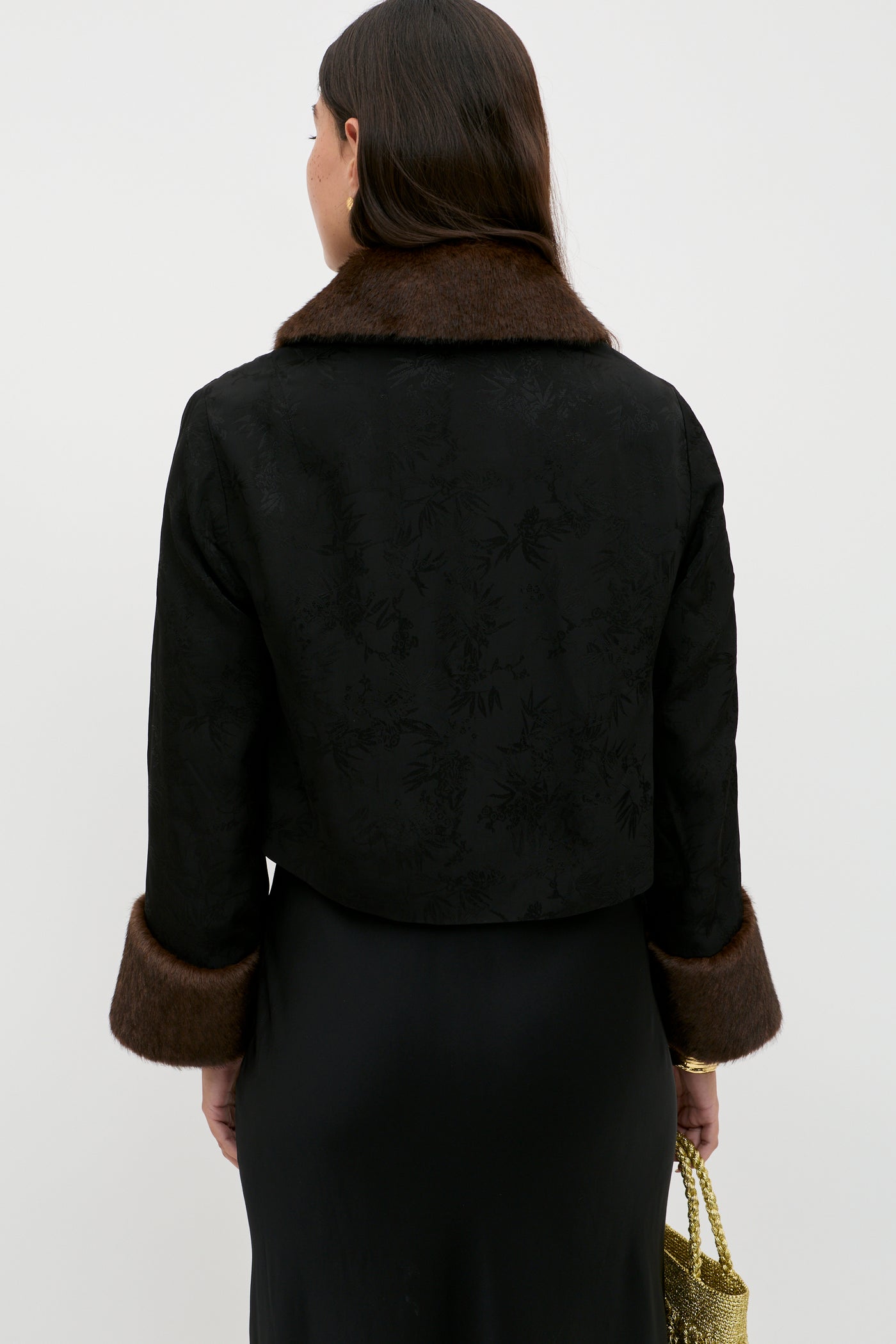 A woman with long brown hair, seen from the back, wears the Luanne - Black jacket with a subtle floral pattern, brown faux fur trim on the collar and cuffs, paired with a black satin skirt and holding a gold purse in her left hand.