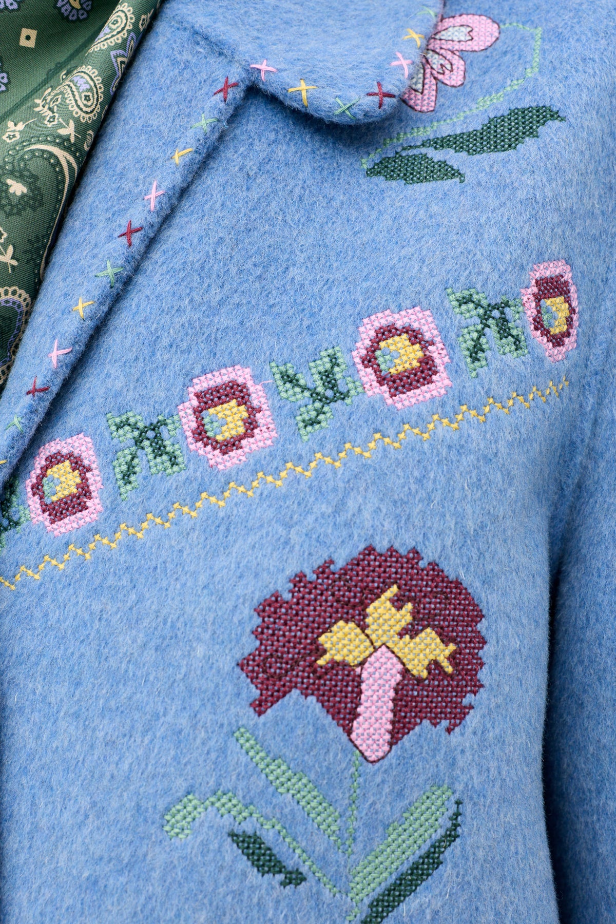 Close-up of the Tashia - Juliette Embroidery Blue wool coat, featuring colorful floral embroidery and cross-stitch detail, worn over a green patterned shirt.