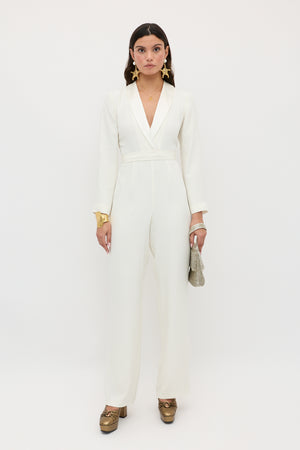 A woman stands against a white background wearing the emilia - ivory jumpsuit with wide-leg trousers, gold shoes, statement earrings, a gold bracelet, and holding a small textured clutch. She looks directly at the camera.