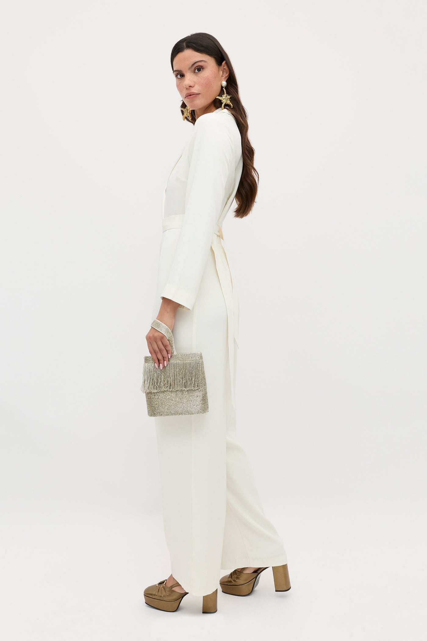 A woman stands against a neutral background, wearing the emilia - ivory seventies-inspired jumpsuit with wide-leg trousers and long sleeves, styled with gold platform shoes, statement earrings, and a silver tasseled clutch.