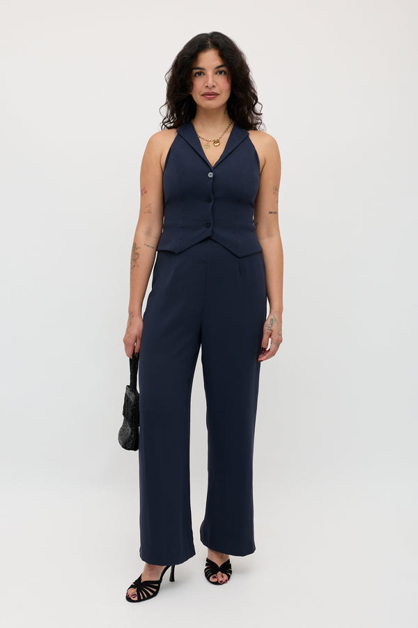 A woman with wavy dark hair and visible arm tattoos stands against a white background, dressed in the Sienna 2 – Navy sleeveless vest and matching pants. She accessorizes with a black handbag and black heels.