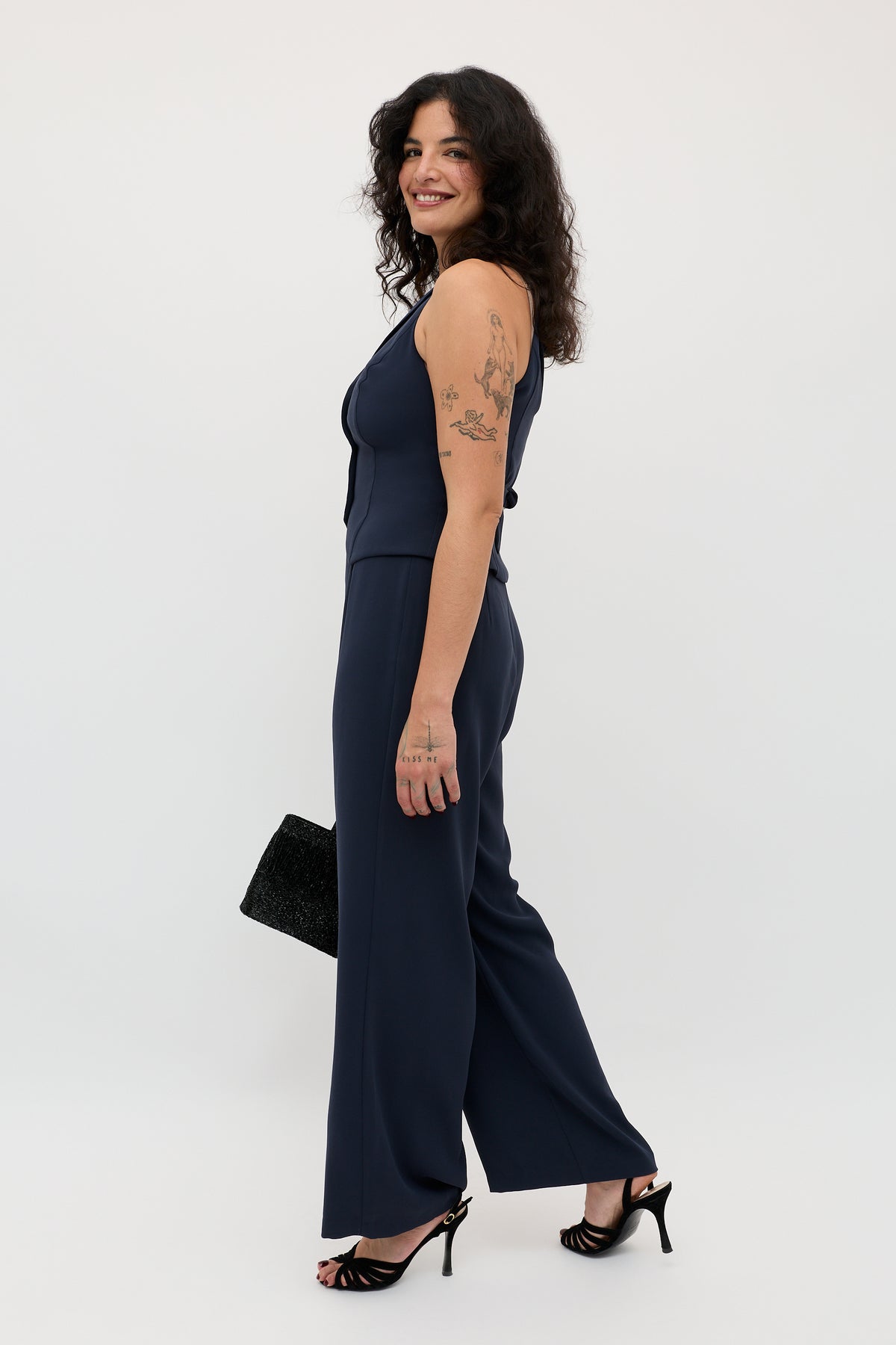 A woman with curly dark hair stands sideways and smiles against a plain white background, wearing the Sienna 2 - Navy sleeveless jumpsuit paired with black heels and holding a black clutch.