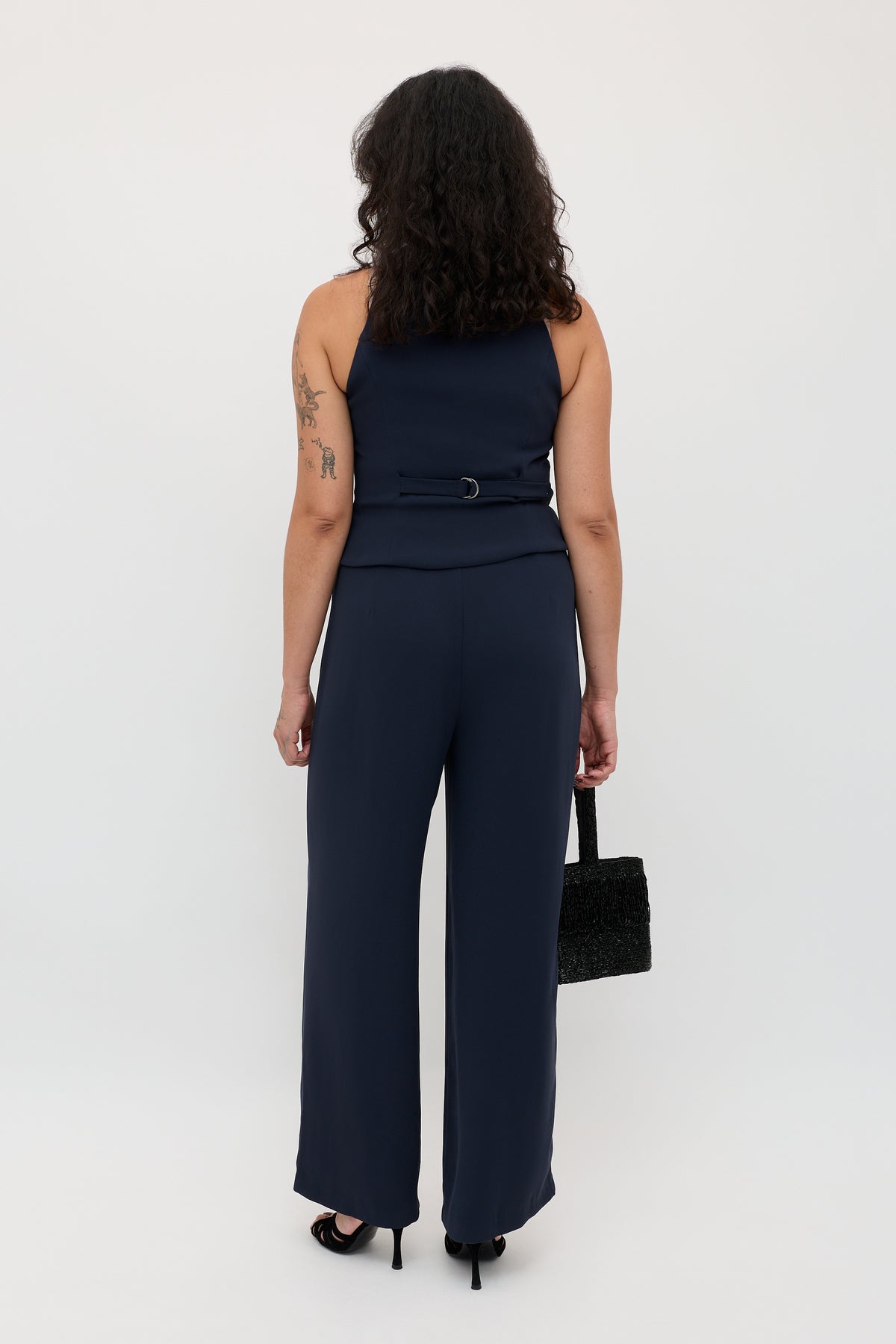 A woman with curly hair and a tattoo on her left arm stands facing away, wearing the Sienna 2 - Navy sleeveless jumpsuit paired with black heels and carrying a black handbag.