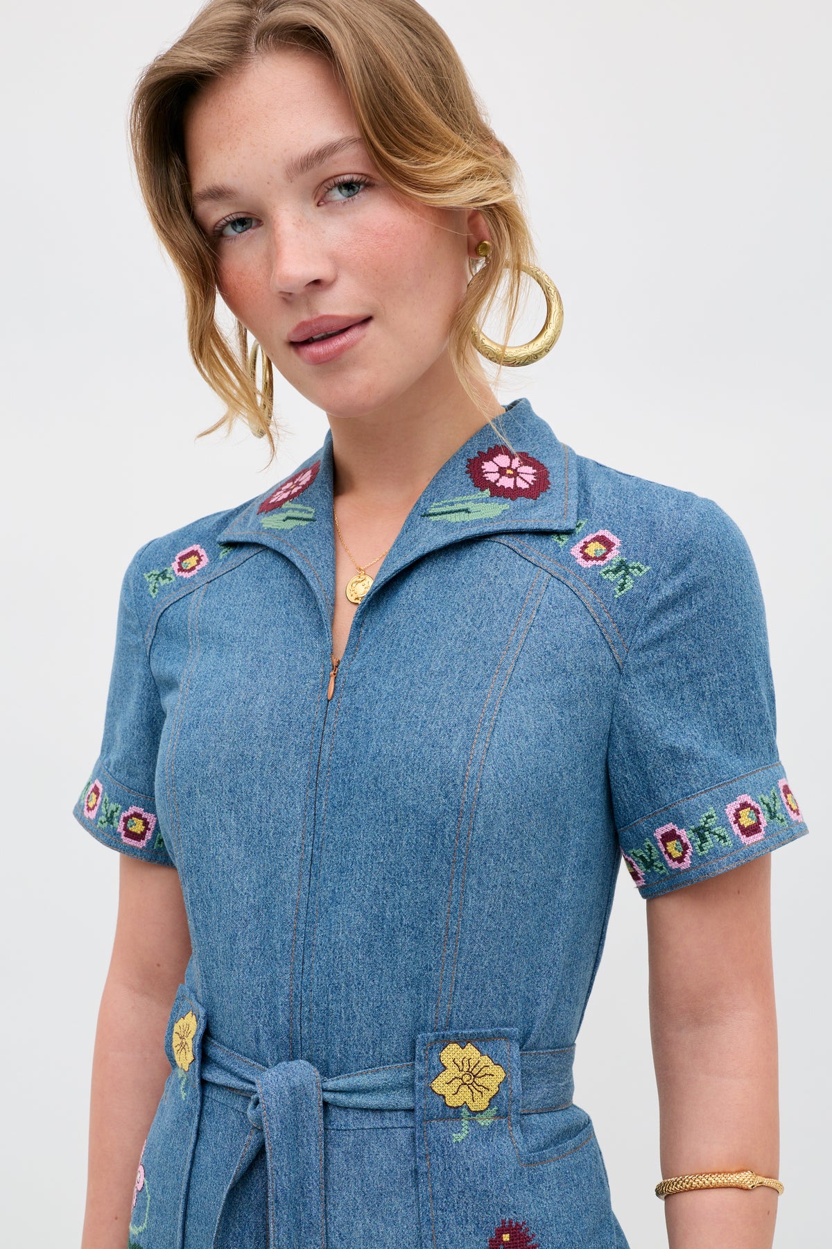 A woman poses against a plain white background wearing the Briana - Juliette Embroidery Blue dress, a blue denim style featuring floral embroidery on the collar, sleeves, and pockets. She has light brown hair and accessorizes with gold hoop earrings and a gold necklace.
