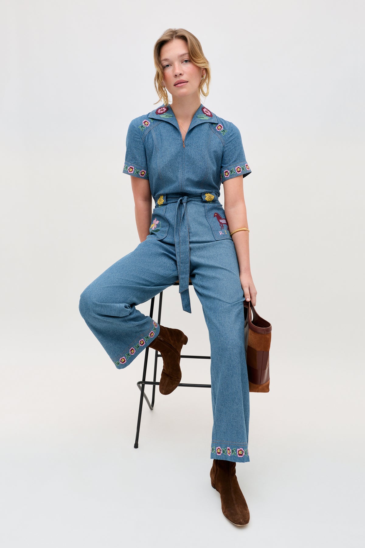 A woman sits on a stool, wearing the Briana - Juliette Embroidery Blue denim jumpsuit with embroidered floral details, paired with brown boots and a brown handbag. The background is plain and light-colored.