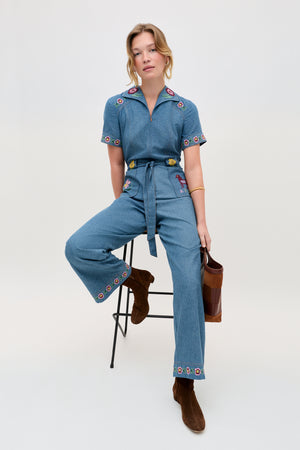 A woman sits on a stool, wearing the Briana - Juliette Embroidery Blue denim jumpsuit with embroidered floral details, paired with brown boots and a brown handbag. The background is plain and light-colored.