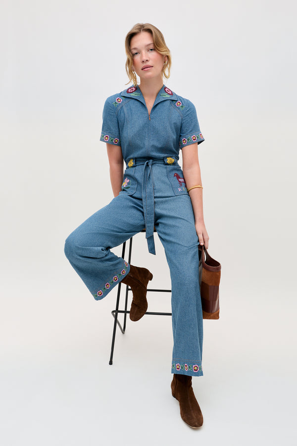 A woman sits on a stool, wearing the Briana - Juliette Embroidery Blue denim jumpsuit with embroidered floral details, paired with brown boots and a brown handbag. The background is plain and light-colored.
