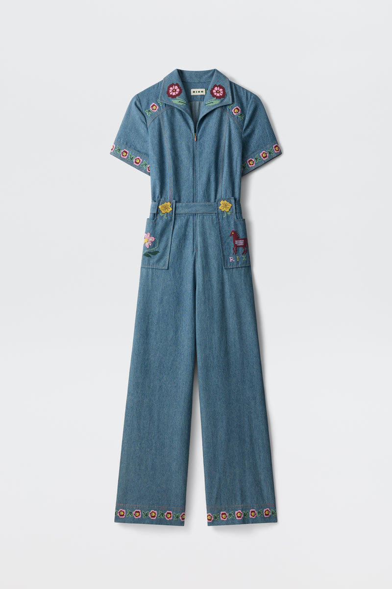 A blue denim jumpsuit, the Briana - Juliette Embroidery Blue, featuring short sleeves and wide legs with colorful floral embroidery on the collar, sleeves, pockets, and hem. One pocket is adorned with an embroidered deer. The jumpsuit is displayed against a plain white background.