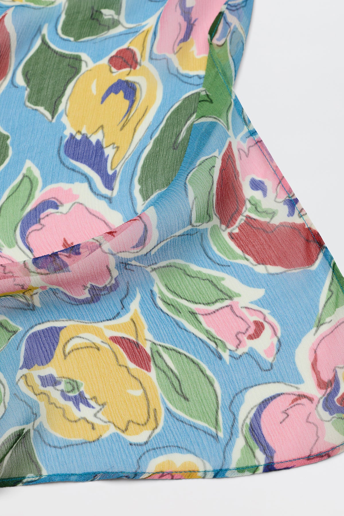 A close-up of Julien - Jeannie Bloom Sky, a sheer, textured fabric with a colorful floral print of pink, yellow, red, and purple flowers and green leaves on a light blue background.