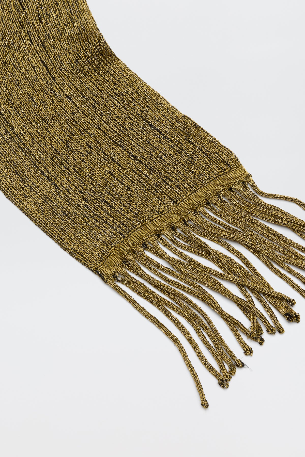 A close-up of the Channing - Gold knit scarf with long, twisted fringe ends, laid flat on a white surface—a glamorous accessory to add shine to any look.