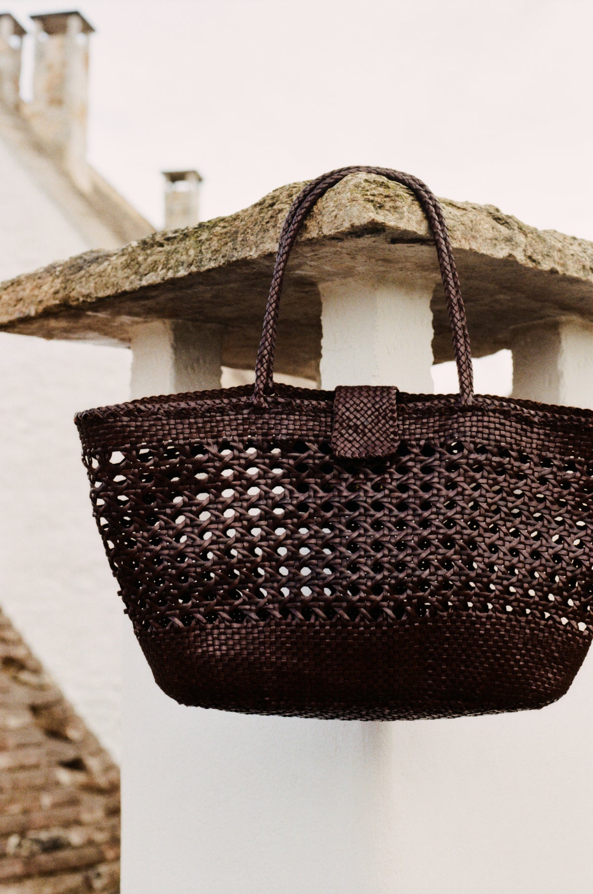 The Lucine - Dark Brown leather woven bag by Dragon Diffusion, featuring two handles, hangs on a white stone pillar with a blurred building and chimney in the background.
