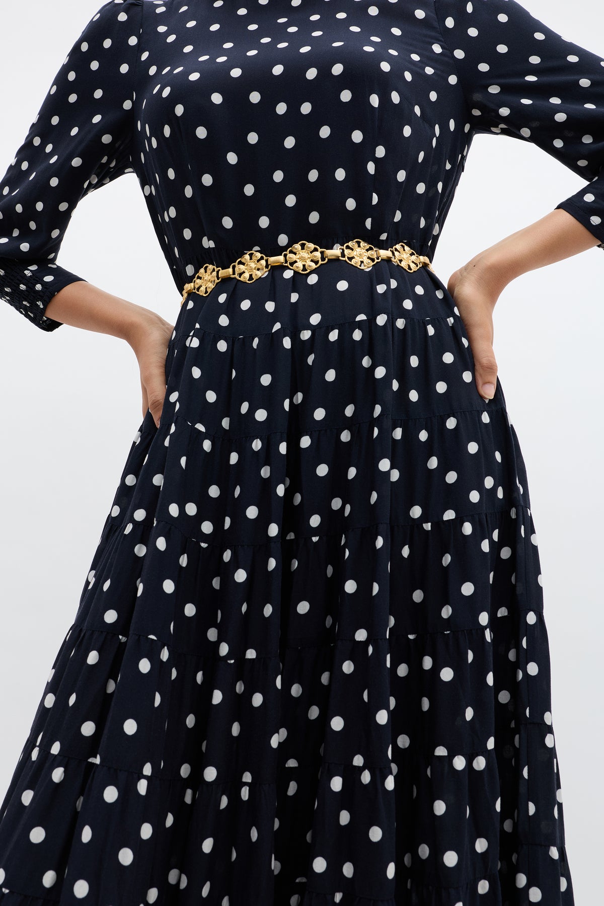 A person wearing a navy blue RIXO dress with white polka dots and the Tiana - Gold daisy-chain belt stands with hands on hips against a plain background.
