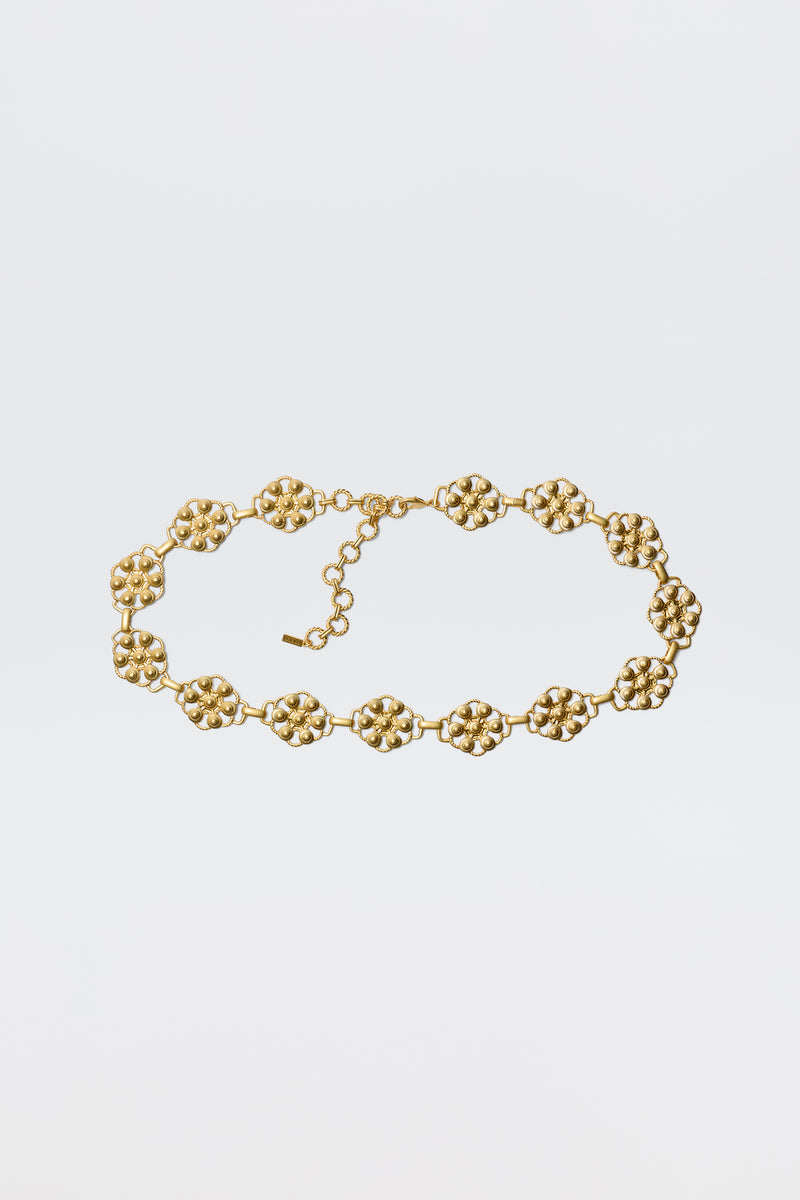 The Tiana - Gold necklace showcases vintage-gold plating with linked, textured floral motifs and an adjustable chain clasp, styled like a daisy-chain belt and displayed against a plain white background.