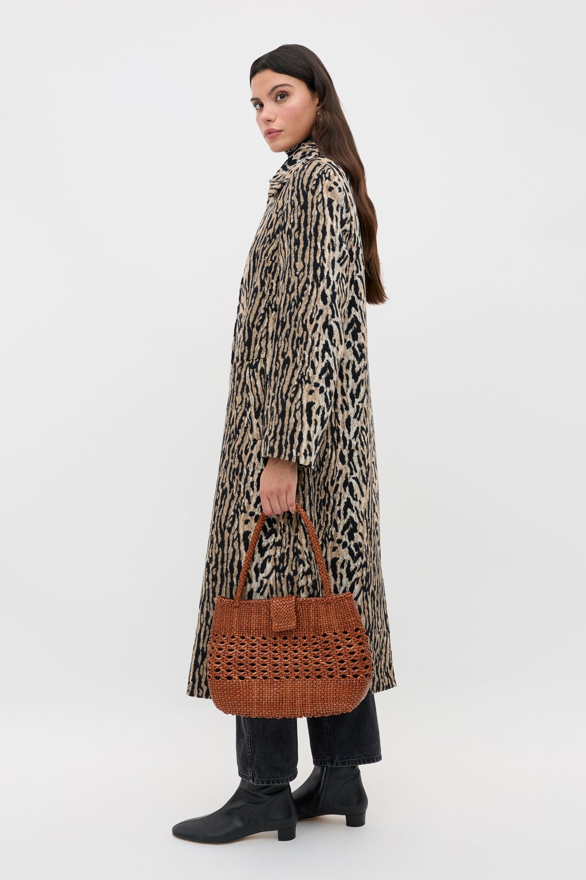 A woman wears the Milly - Bohemia Leopard mid-length coat, paired with black pants and boots, holding a brown woven handbag as she stands against a plain white background and glances over her shoulder.