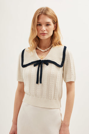 A woman with light brown hair wears the Pippa - Ivory: a vintage-style cream nautical knit top with short sleeves, a large square collar trimmed in black, a black ribbon tie, paired with a light skirt and layered necklaces.