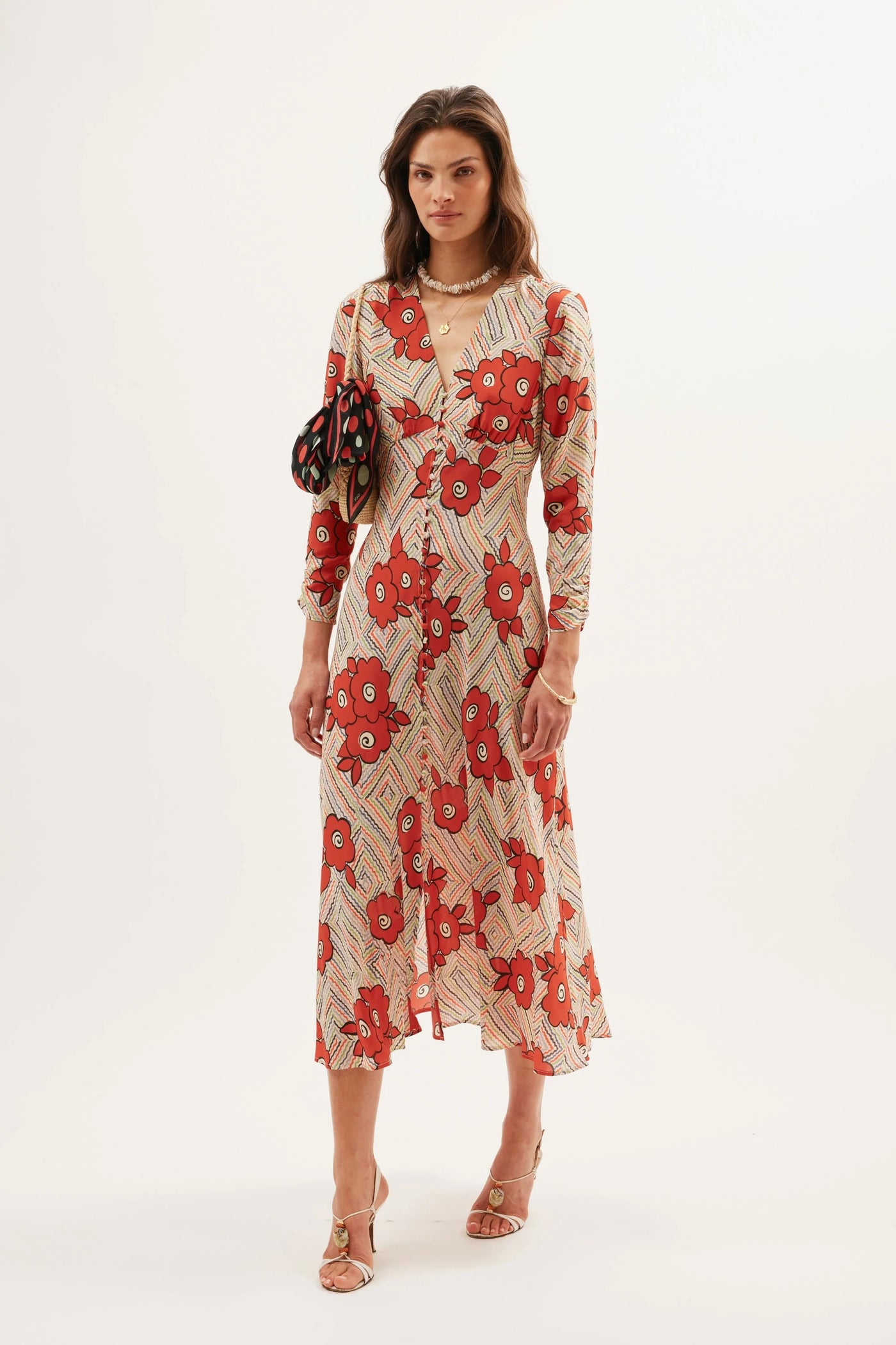A woman poses in the Katie - Milos Floral Cream dress featuring long sleeves and a red-beige floral print. She holds a black handbag with circular accents, wears strappy sandals, gold jewelry, and stands against a plain white background.