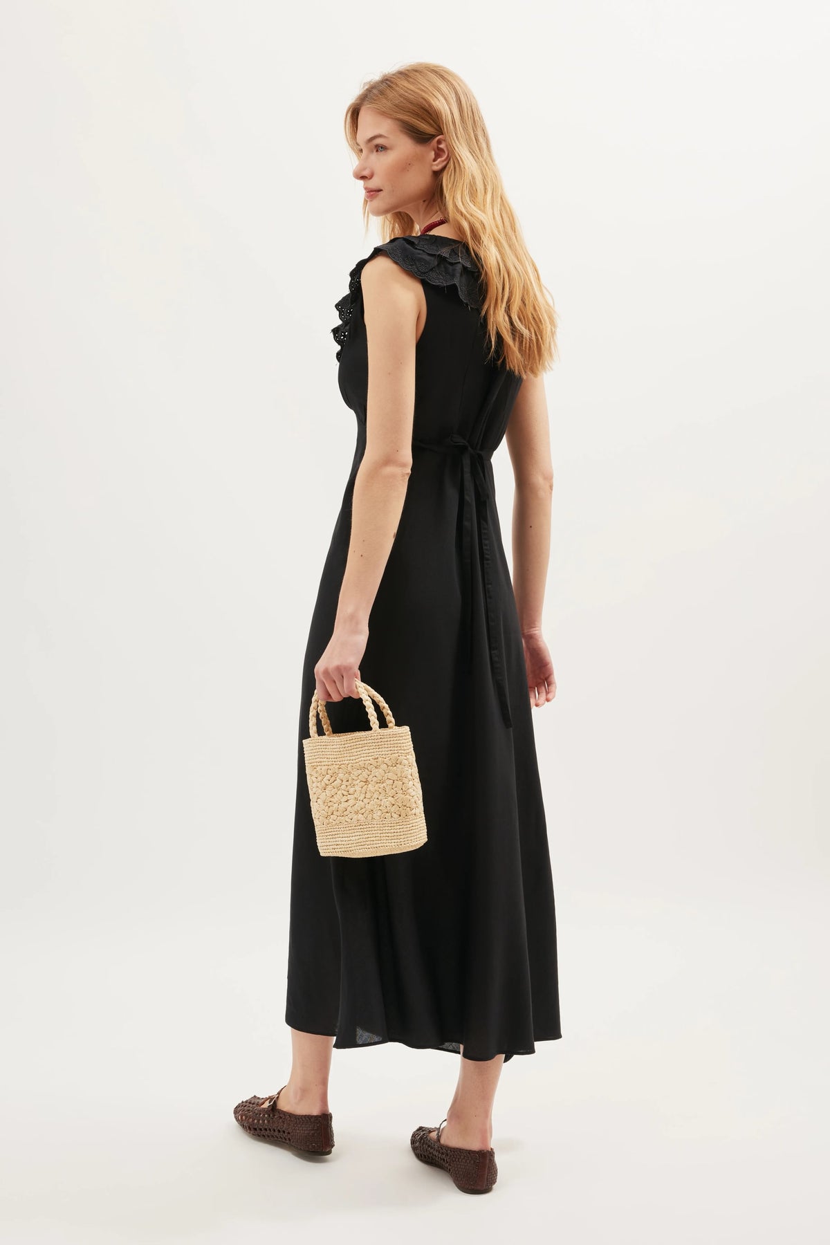A woman with long blonde hair wears the Seraphina - Black dress, a sleeveless belted maxi featuring scalloped ruffles, holding a small straw handbag and standing slightly turned to the side against a plain white background.