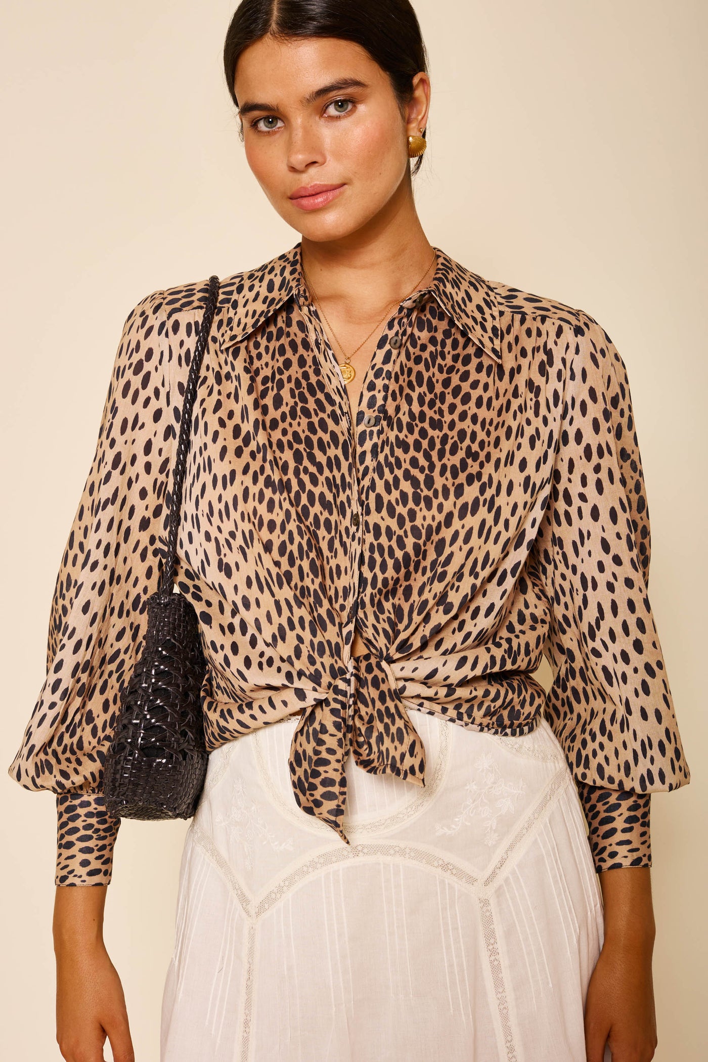 A woman wears the Blake - Antelope Spot vintage animal print shirt tied at the waist, paired with a white lace skirt, gold earrings, a necklace, and a black textured shoulder bag against a neutral background.