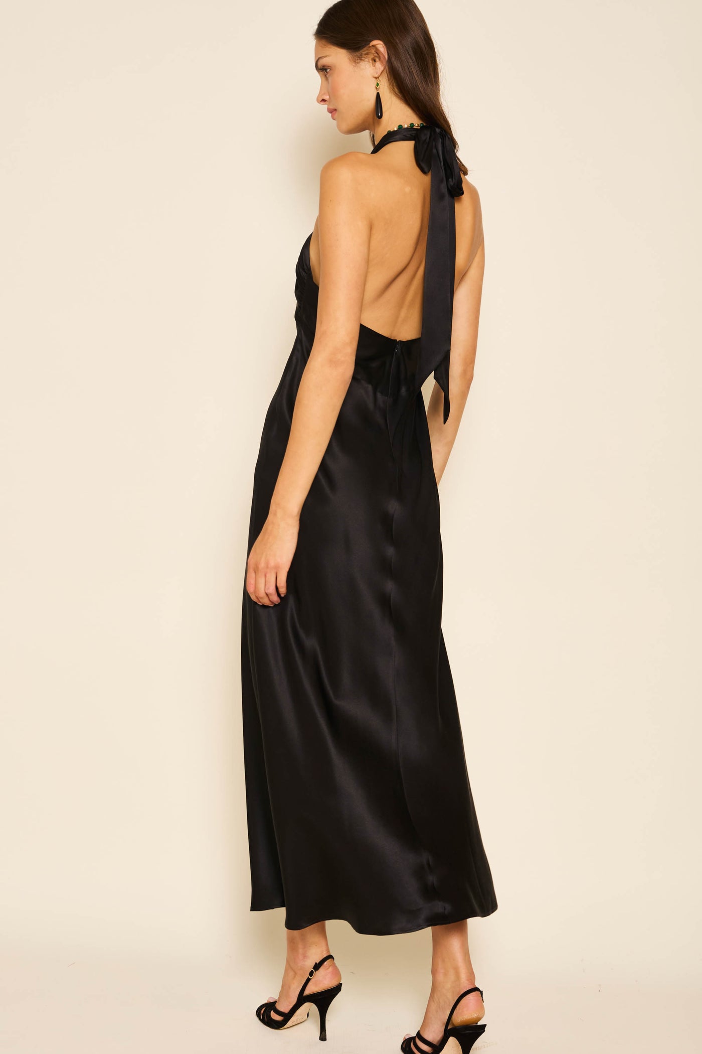 A woman stands sideways, looking down, in the Dune - Black long silk-satin halterneck gown with an open back and bow detail. She wears black high-heeled sandals, drop earrings, and has dark hair.