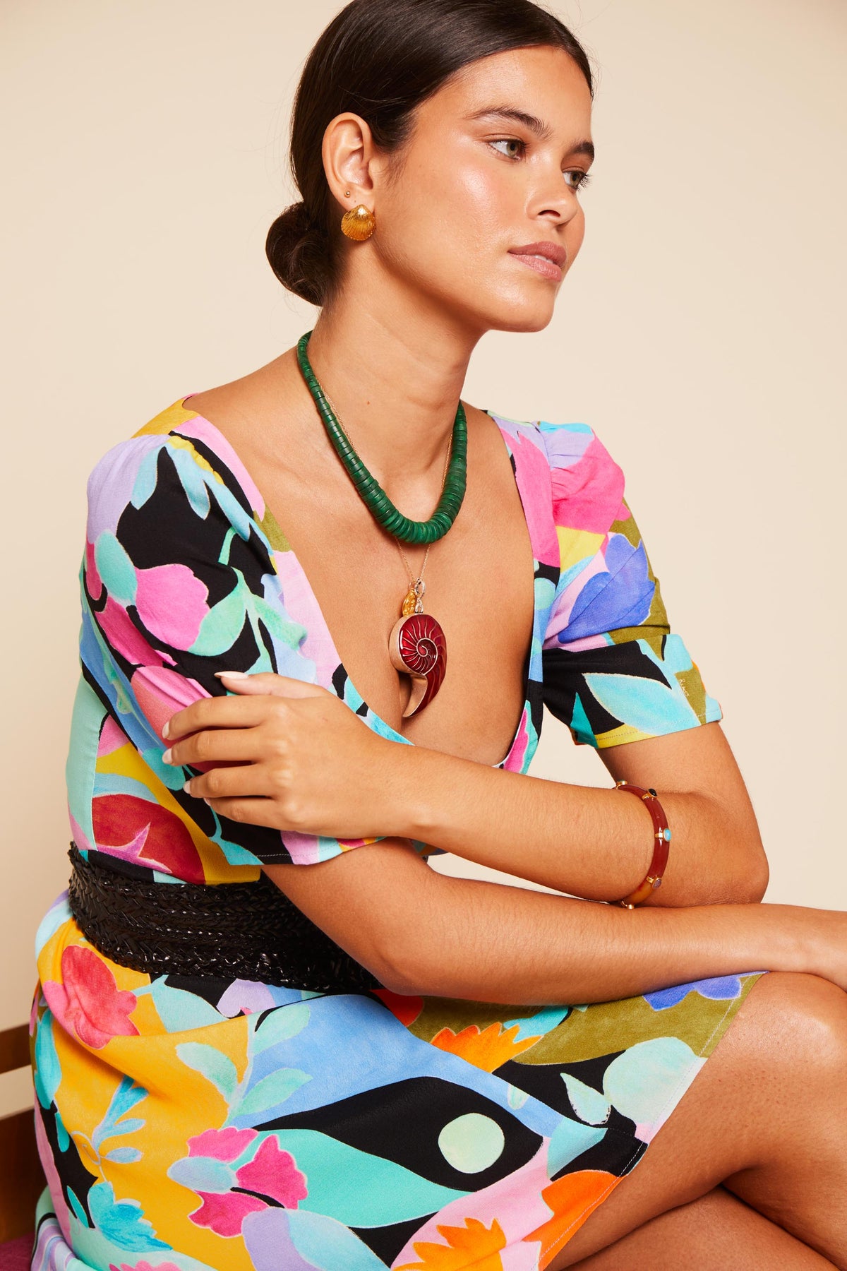 A woman in a colorful floral dress sits with arms crossed, wearing vintage earrings, a green beaded necklace with a large pendant, a brown bangle, and the Meyra - Gold. Her hair is pulled back as she gazes thoughtfully to the side.