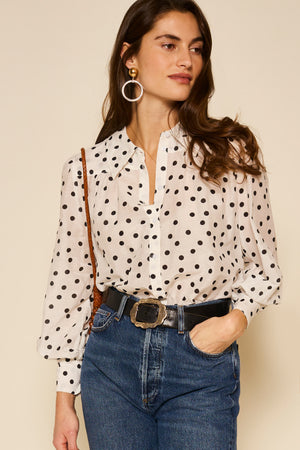 A woman wearing the Blake - Bohemia Spot Top, blue jeans, a black belt with a large buckle, hoop earrings, and a brown shoulder bag stands against a beige background.