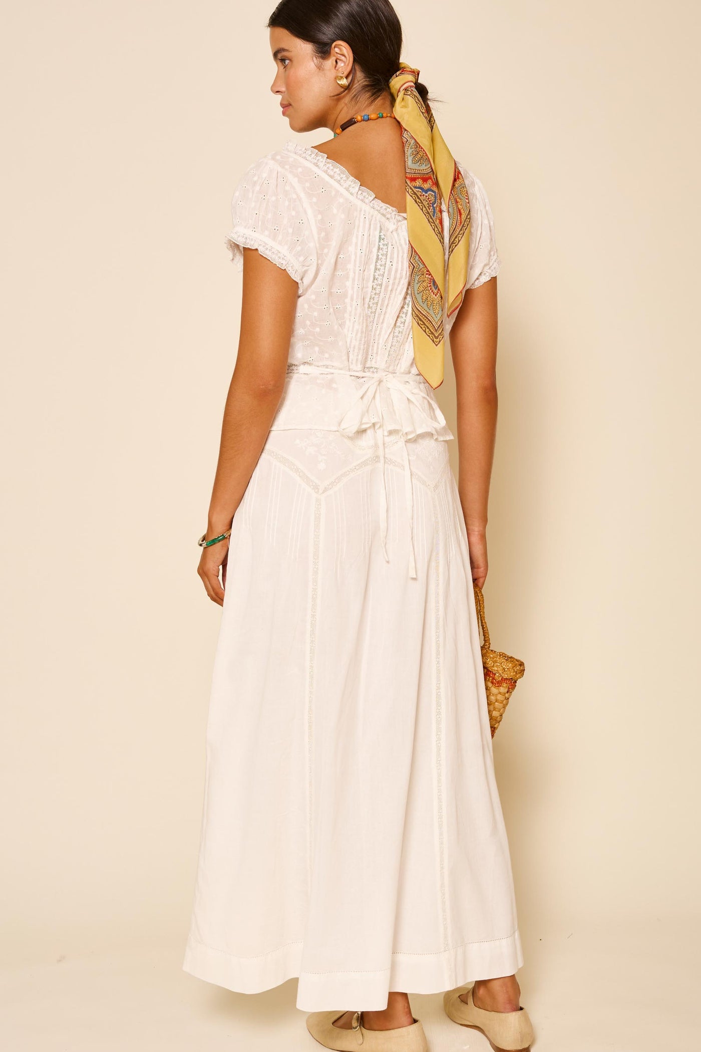 A woman, facing away against a beige background, showcases the Vienne – Pansy Embroidery Ivory dress with lace details and a back bow. She accessorizes with a yellow patterned hair scarf, neutral shoes, and carries a woven bag.