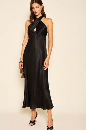 A woman in the Dune - Black sleeveless silk-satin gown with a crisscross halter neckline and front cutout, accessorized with a gold clutch and black heels, stands before a beige backdrop—ideal for any black tie dress event.
