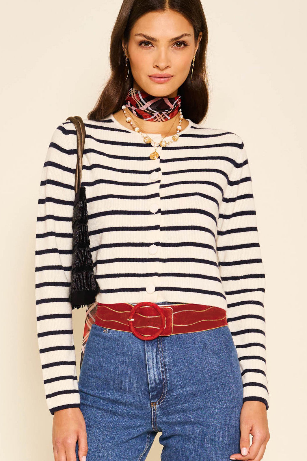 A woman stands against a beige background wearing a black and white striped sweater, blue jeans, a patterned neck scarf, and a shoulder bag with black tassels, accessorized with the Catalina - Red suede belt.