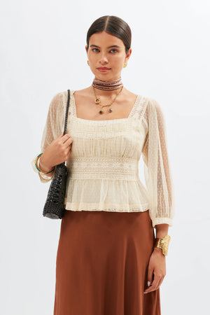 A woman wearing the Tyla - Vintage Cream blouse with sheer Swiss dot tulle sleeves, paired with a brown skirt and gold jewelry, stands against a plain white background carrying a black woven bag.