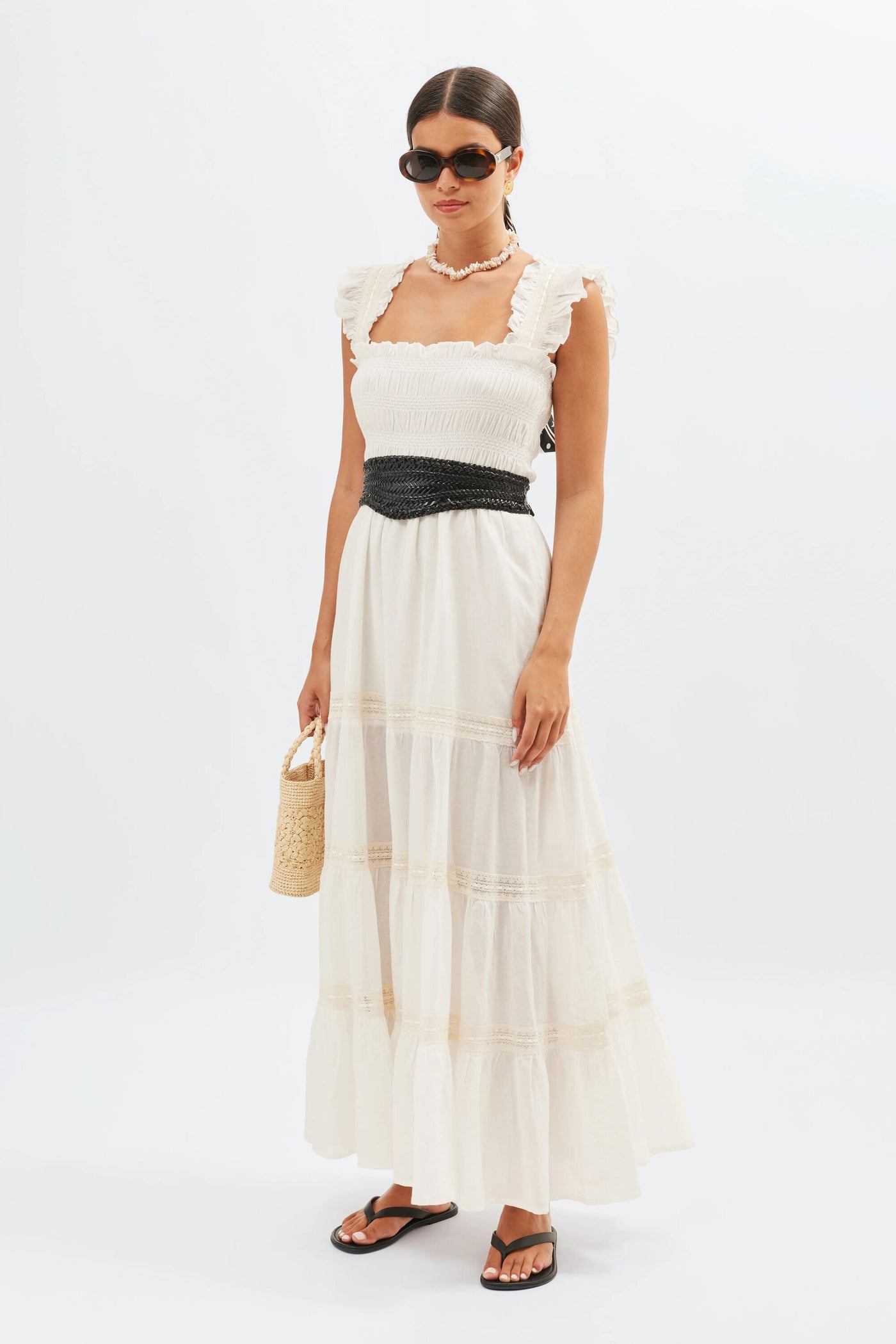 A woman wearing the Aubrie - Ivory white, ruffled nightdress styled as a maxi dress, cinched with a black belt and paired with black sandals and sunglasses, holds a small woven bag while standing against a plain white background.