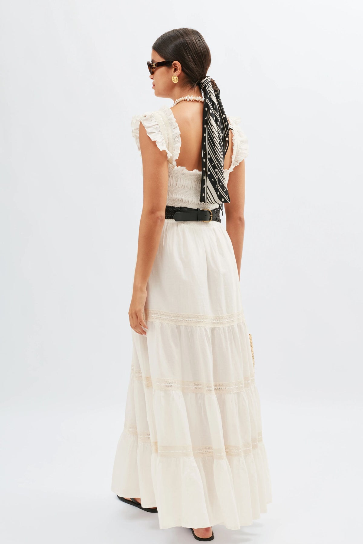 A woman wearing the Aubrie - Ivory, a long, white, 70s-inspired nightdress with ruffled straps, stands facing away. She has a black and white scarf tied in her hair, sunglasses, gold earrings, a black belt, and black sandals.