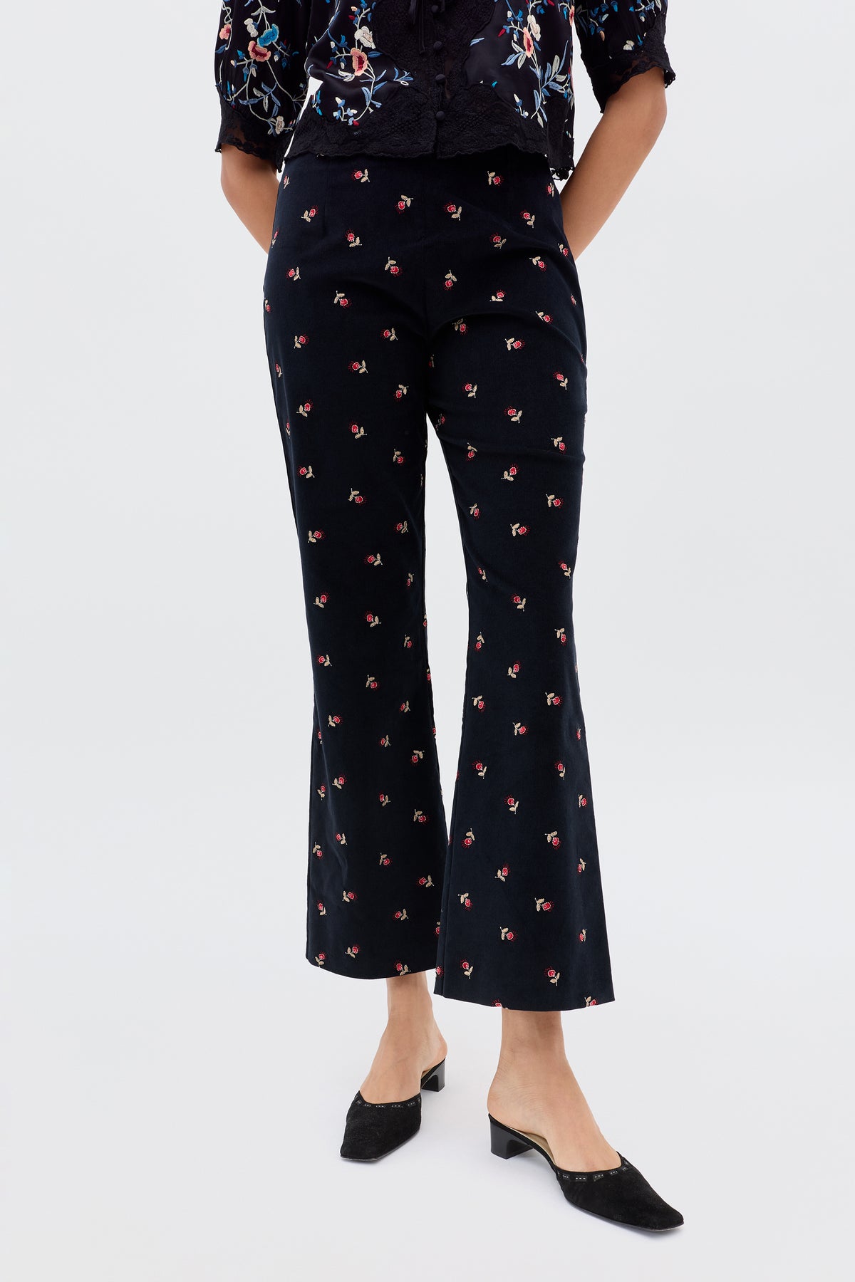 A person wears Olwen - Dandy Embroidery Deep Navy trousers, featuring a black base with small pink floral embroidery, paired with black low-heeled shoes against a plain light background. The upper body is partially visible.