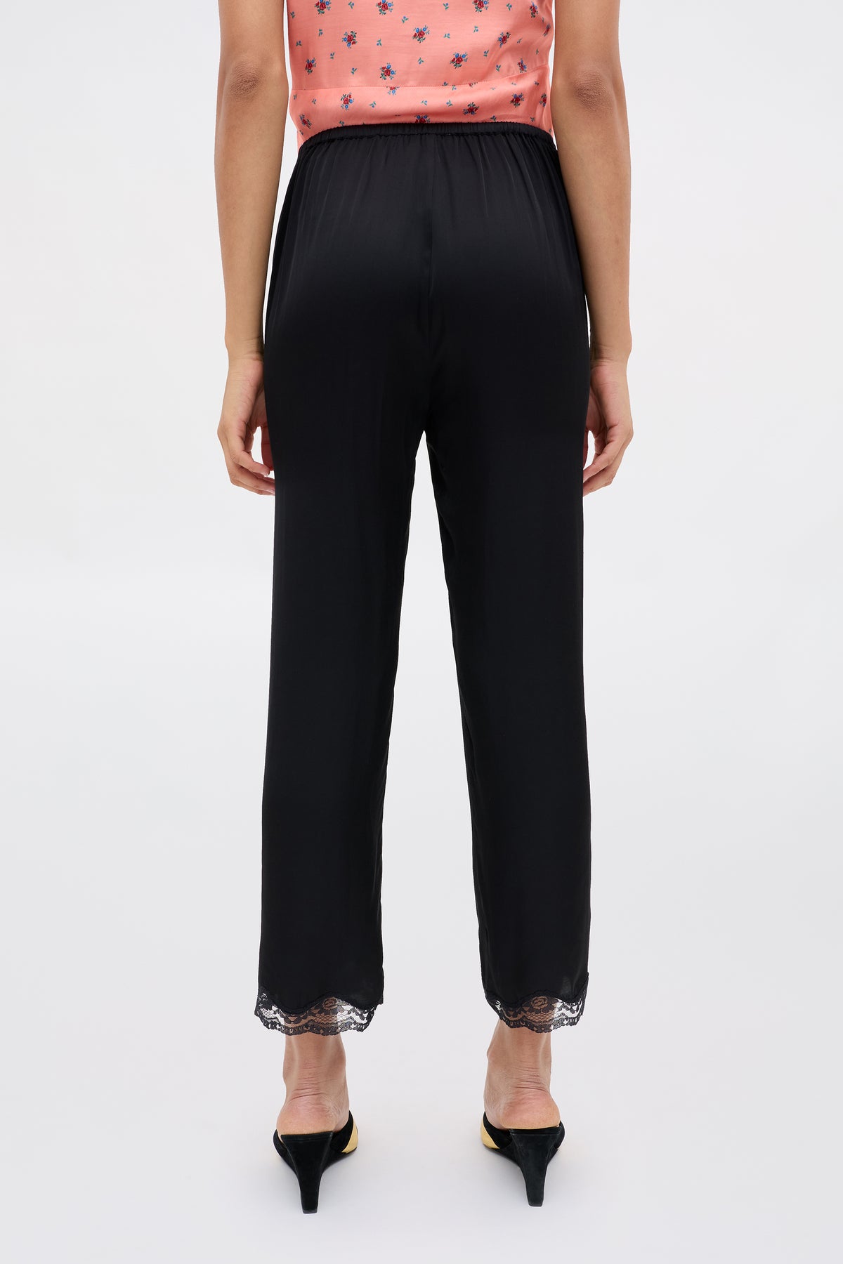 A person is shown from the back wearing Mireille - Black high-waisted satin pants with lace trim at the ankles and black heels, paired with a peach sleeveless floral top.