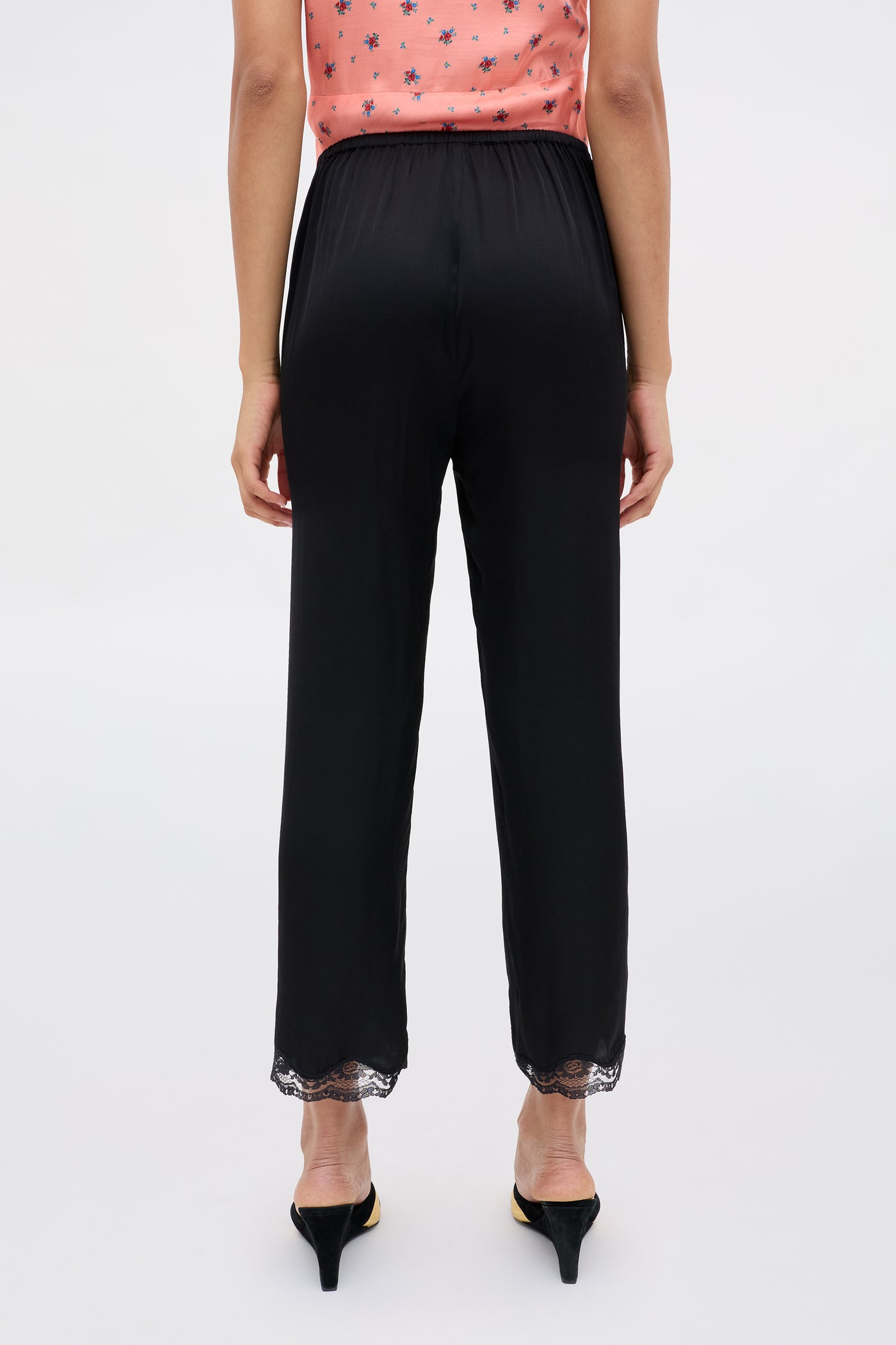 A person is shown from the back wearing Mireille - Black high-waisted satin pants with lace trim at the ankles and black heels, paired with a peach sleeveless floral top.