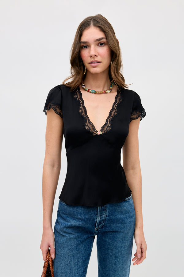 A woman wearing the Karia - Black lace-trimmed V-neck top, blue jeans, and a colorful beaded necklace stands against a plain light background, holding a brown bag.