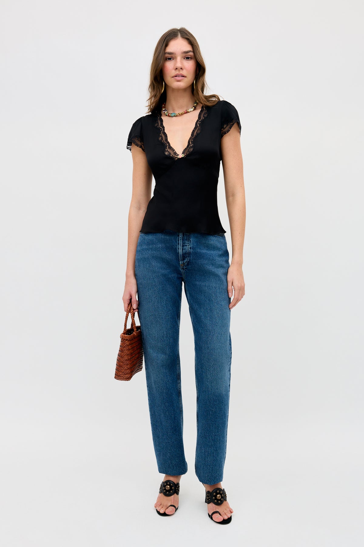 A woman stands against a plain background wearing the Karia - Black lace-trimmed top, blue jeans, black sandals, a beaded necklace, and holding a small brown woven handbag.