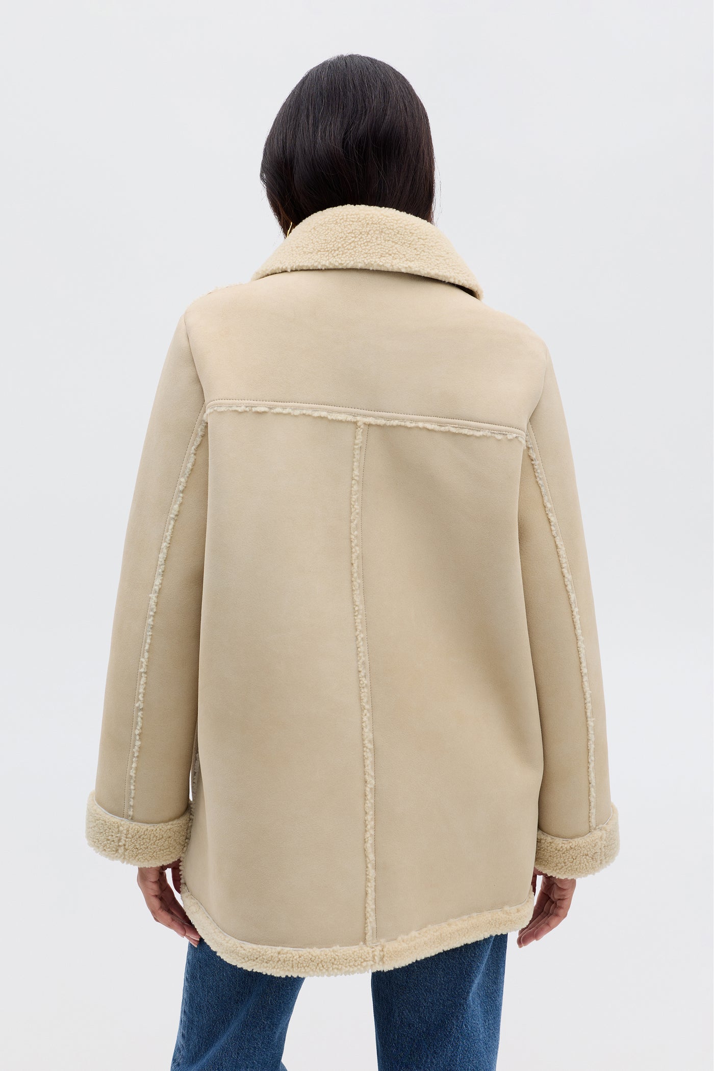 A person faces away, wearing Ellis - Cream: a beige faux-shearling coat with visible seams and a wide collar, paired with blue jeans against a plain, light background.