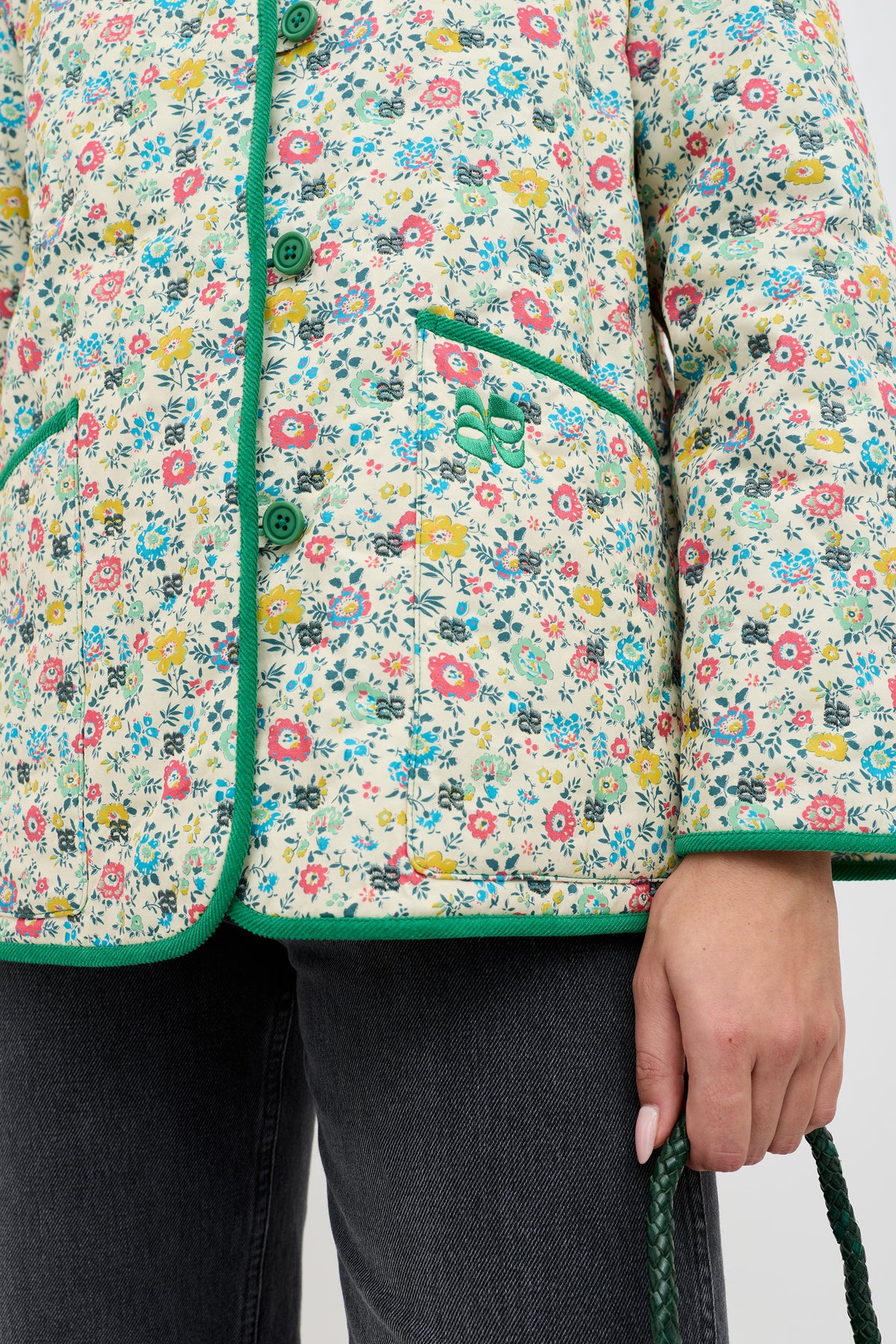 Wearing the Earla - Amalfi Floral Cream jacket with green trim and buttons, paired with black jeans and holding a green braided strap. Only the torso and one hand are visible.